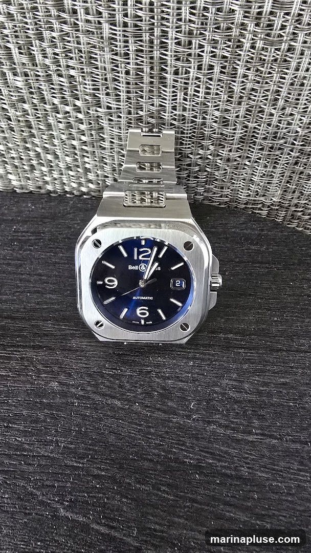 A close-up shot of a Bell & Ross watch with a dark blue dial and a stainless steel bracelet. The watch is placed on a dark, textured surface, with a metallic mesh background. The watch face displays Arabic numerals at 12, 6, and 9, with white indices for other hours. The brand name "Bell & Ross" is visible below the 12 o'clock position, and "AUTOMATIC" is printed below the center. A date window is present at the 3 o'clock position.