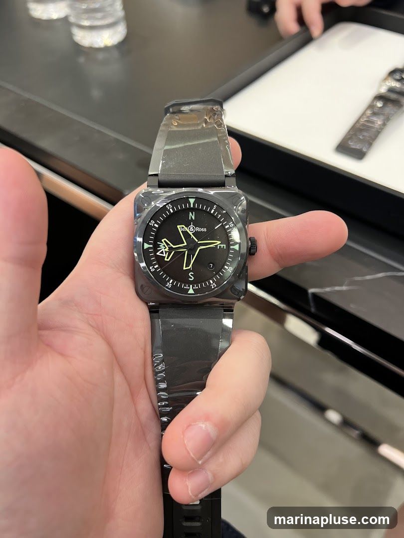 A person is holding a black Bell & Ross watch with a square face. The watch face has a compass rose with N, S, E, and W markings, and a stylized airplane graphic. The watch band is also black. In the background, there are bottles of water and another watch on display.