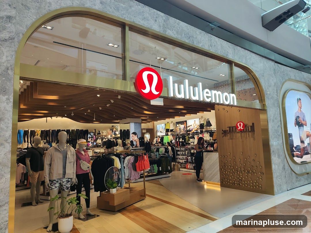 The exterior of a Lululemon store with its prominent logo and name displayed above the entrance. Inside, mannequins are dressed in athletic wear, and racks of clothing are visible. The store features a modern design with wood accents and marble-like finishes.