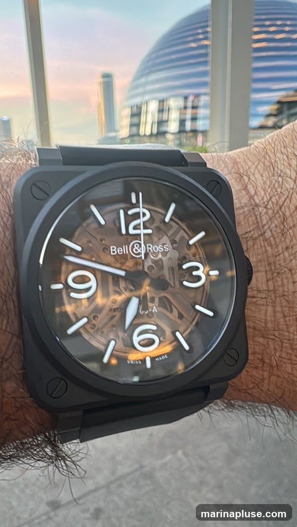 A close-up shot of a Bell & Ross watch on a person's wrist. The watch has a black square case and a skeletonized dial with white Arabic numerals and hands. The background shows a blurred cityscape with a distinctive domed building and a tall skyscraper, under a soft, colorful sky.
