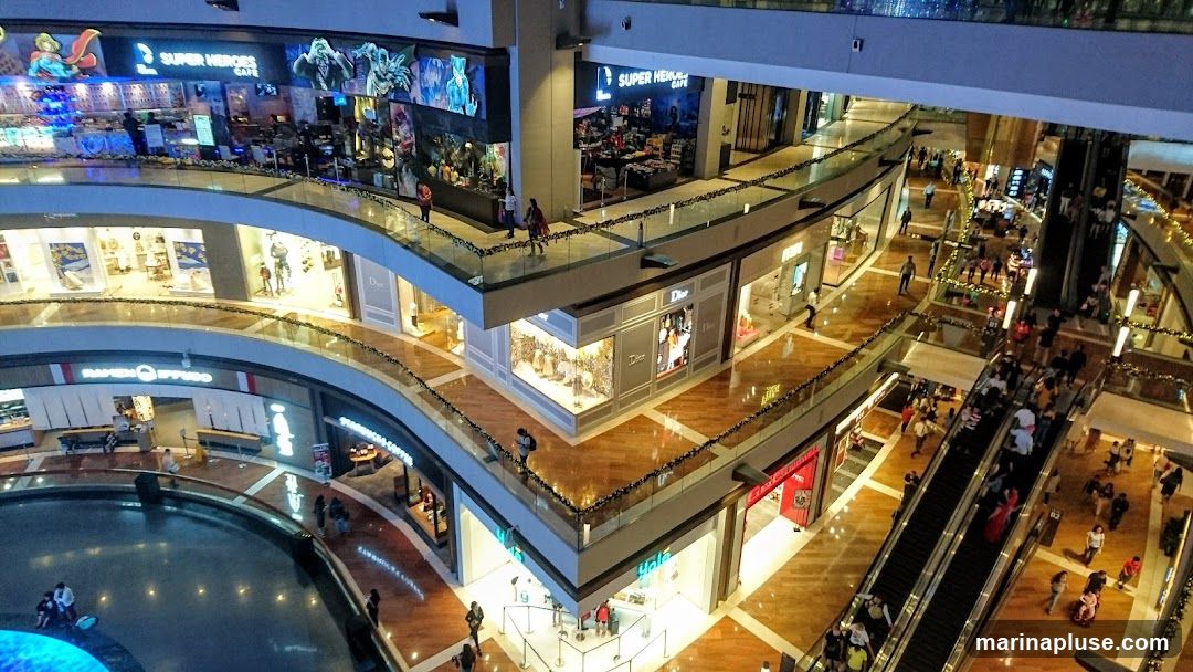 An aerial view of the interior of a large, modern shopping mall. Multiple levels are visible, connected by escalators and walkways. Various shops with illuminated storefronts are present, including Dior and Super Heroes Cafe. People are seen browsing, walking, and using the escalators. The mall features polished wooden floors and decorative lighting.