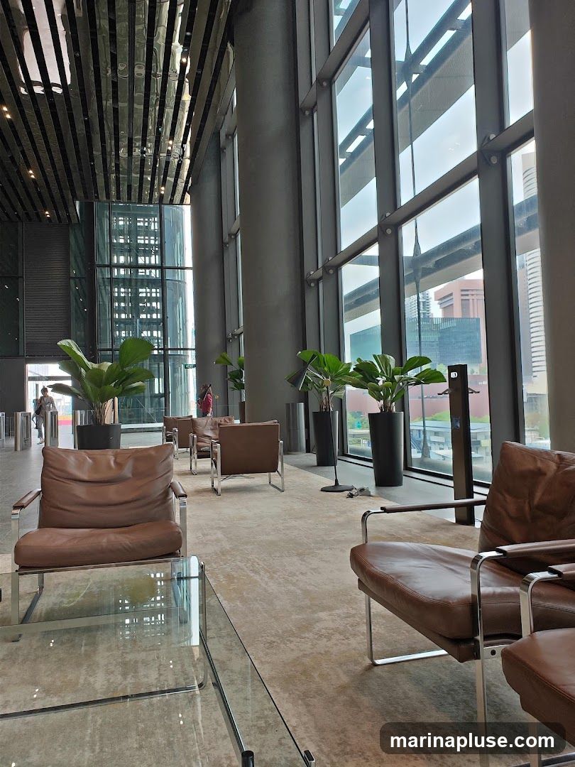 A spacious and modern office lobby with large windows, comfortable leather seating, and potted plants. The interior features a high ceiling with metallic accents and concrete flooring. Natural light streams in through the expansive windows, offering a view of the city skyline.