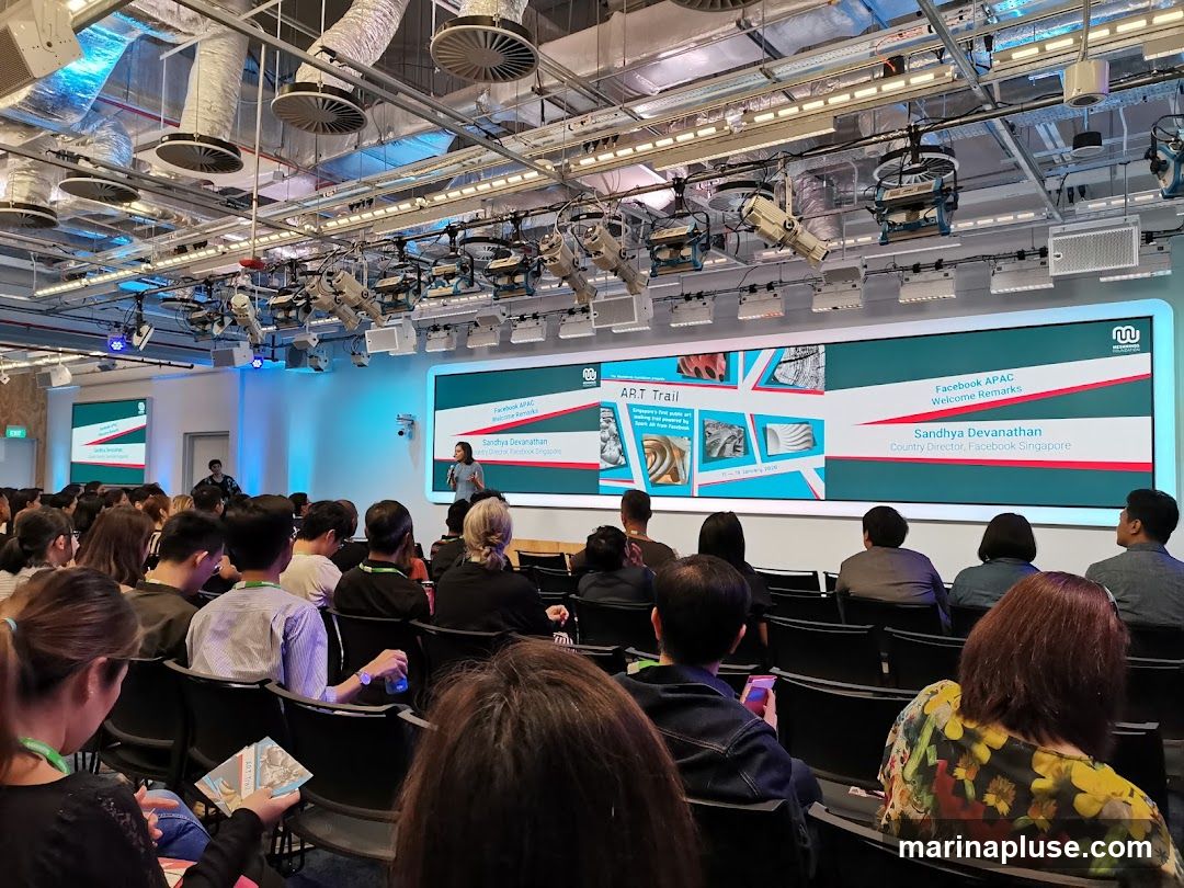 A presentation is taking place in a large room with an audience seated in rows of chairs. A speaker, Sandhya Devanathan, Country Director of Facebook Singapore, is presenting on a large screen. The screen displays information about the 'ART Trail' and 'Facebook APAC Welcome Remarks'. The room has industrial-style lighting and ventilation systems visible on the ceiling.