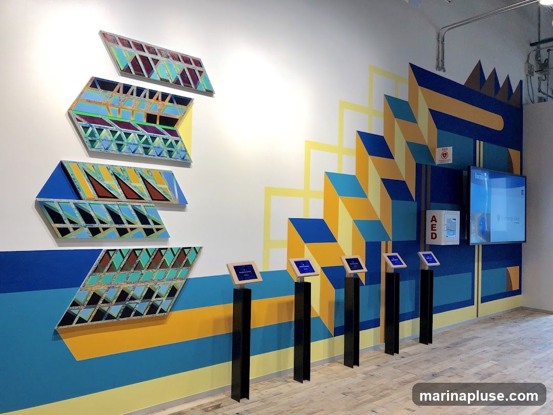 The image shows a brightly lit interior space with a large, colorful geometric mural on the wall. Several abstract art pieces, also featuring geometric patterns, are mounted on the mural. In front of the mural, there are four digital display screens mounted on pedestals, and to the right, a larger screen displays the Facebook logo and text that reads 'Community Voices'. Two AED (Automated External Defibrillator) boxes are also visible on the wall. The floor is made of light-colored wood planks.
