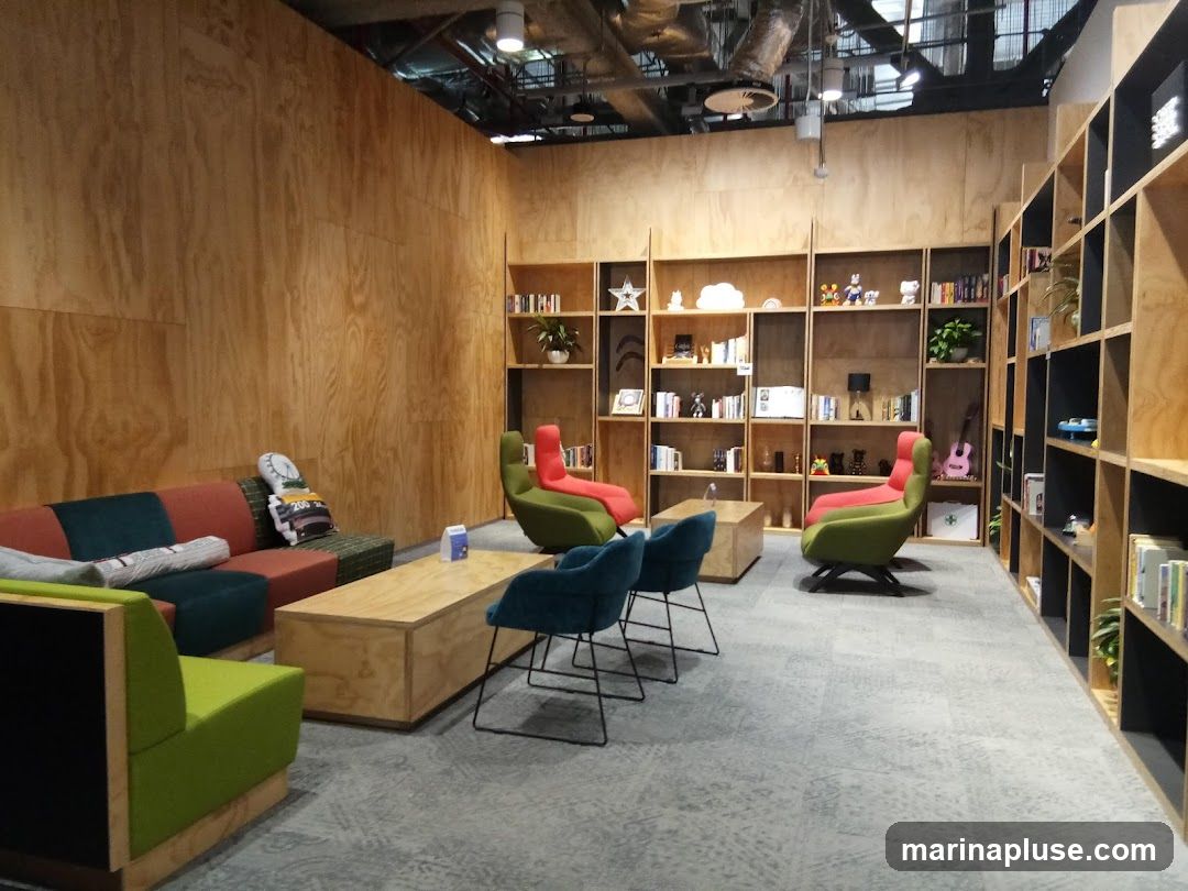 A contemporary lounge area with wooden paneled walls and floor-to-ceiling bookshelves. The furniture includes a modular sofa in shades of red and teal, a green accent chair, and two teal chairs around a wooden coffee table. The bookshelves are filled with books, decorative items, and a guitar.