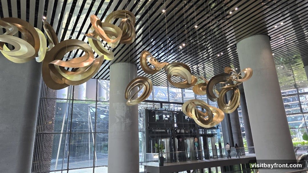 A large, abstract sculpture made of metallic, curved shapes hangs from the ceiling in a modern building lobby. The sculpture is gold and bronze in color and is suspended by thin wires. The lobby features large windows with a view of the exterior, columns, and a reflective ceiling.