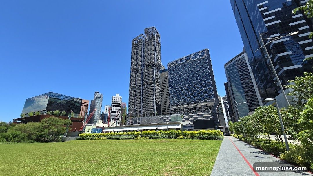 A wide-angle view of a modern cityscape under a clear blue sky. In the foreground, a lush green lawn stretches out, bordered by a paved walkway with a red line. Beyond the lawn, a row of manicured bushes and trees lines the walkway. The background is dominated by a cluster of contemporary skyscrapers, some with unique architectural designs, including one with a honeycomb-like facade and another with a more intricate, layered structure. A glass-fronted building with a distinctive angular design is also visible. The scene suggests a well-developed urban area with integrated green spaces.