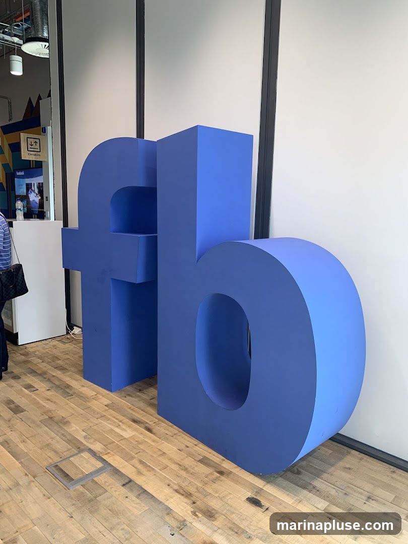 A large, blue, three-dimensional sculpture of the Facebook logo is displayed indoors. The sculpture is positioned on a wooden floor in front of a white wall. In the background, there are signs and a glimpse of people.
