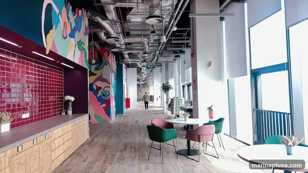 A long, modern office hallway with a vibrant mural on the left wall. The mural features abstract colorful shapes and figures. To the left of the mural is a counter with red tiled backsplash and wooden cabinets below. The hallway has exposed ceiling pipes and ventilation systems. On the right side, large windows let in natural light, with white blinds partially covering them. Several round tables with colorful chairs are placed in the seating area. A person is walking away from the camera down the hallway, towards an "EXIT" sign.