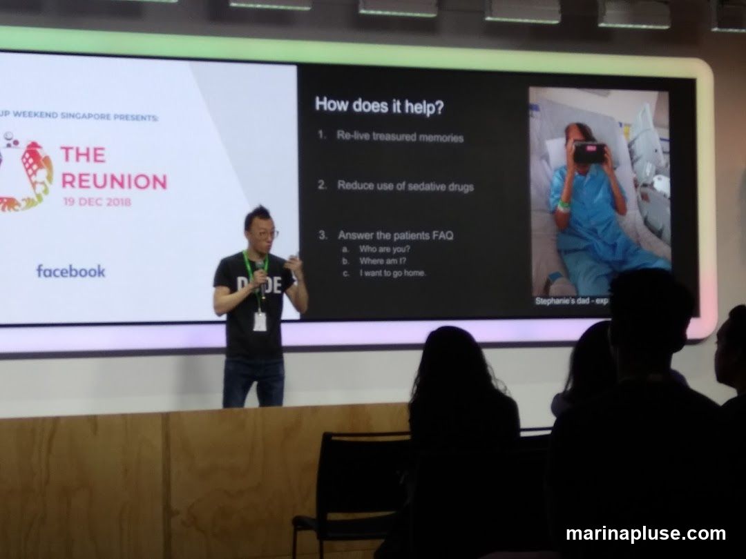 A man in a black t-shirt with 'DE' printed on it is giving a presentation on a large screen. The screen displays text about a "Reunion" event on "19 DEC 2018" presented by "Startup Weekend Singapore" and sponsored by "facebook". Another section of the screen asks "How does it help?" and lists three points: "Re-live treasured memories", "Reduce use of sedative drugs", and "Answer the patients FAQ" with sub-points about identity and location. An image shows a patient in a hospital bed wearing a VR headset. The audience is silhouetted in the foreground.