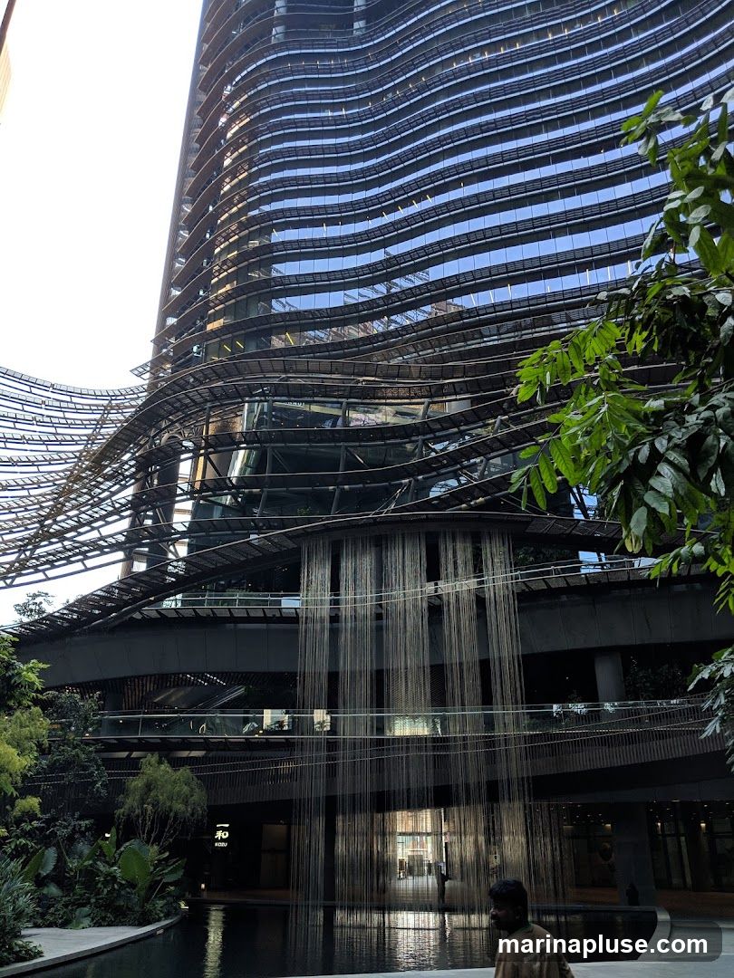 A man walks past a reflective pool in front of a modern, multi-story building with a distinctive wavy facade. The building features extensive glass windows and a unique architectural design with suspended elements and what appears to be a water curtain cascading down. Lush greenery surrounds the area, adding to the ambiance.