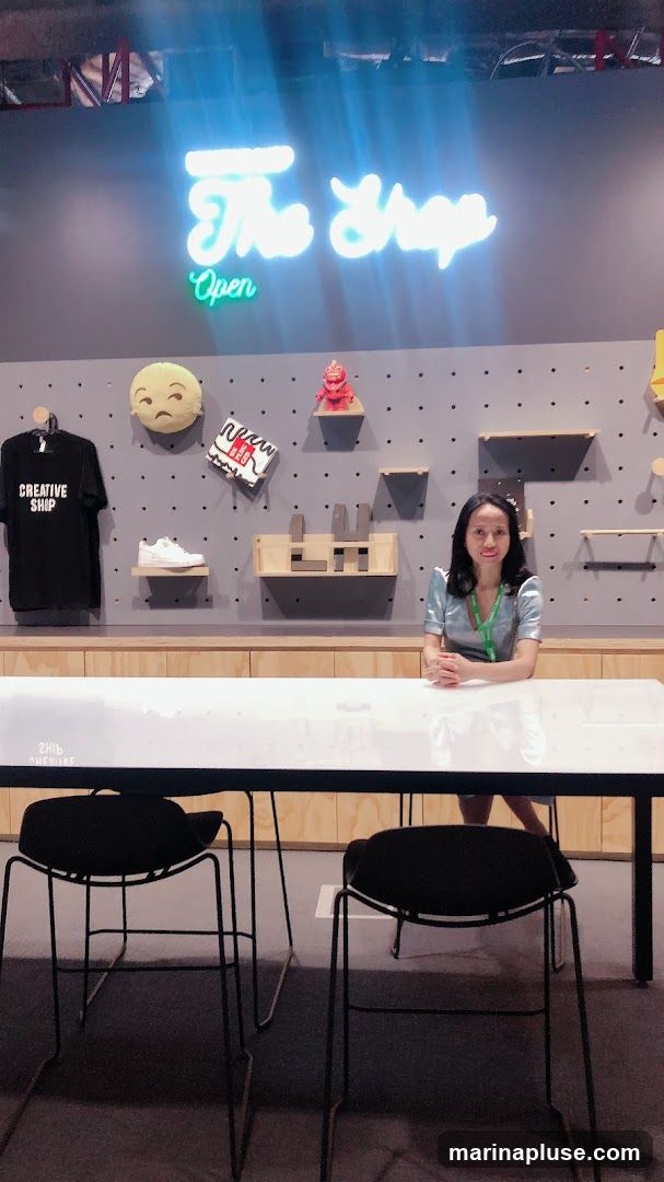 A woman sits behind a long white table in a store. Above her, a neon sign reads "The Shop" and "Open". The wall behind her is grey with a pegboard design, displaying various items including a black t-shirt with "CREATIVE SHOP" printed on it, a white sneaker, a plush emoji pillow, and small decorative figures.