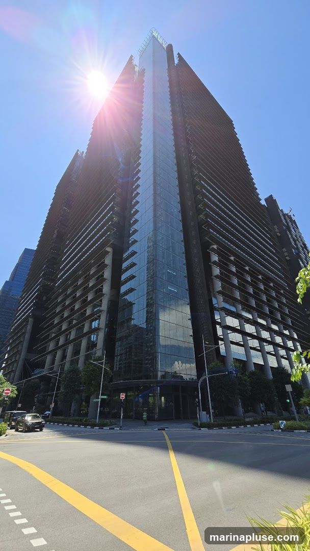 A low-angle shot of a modern, dark-colored skyscraper with a glass facade, set against a clear blue sky. The sun is shining brightly, creating lens flare. The building has a distinctive stepped design with horizontal elements. The street in front of the building is visible, with yellow lane markings and a few cars.