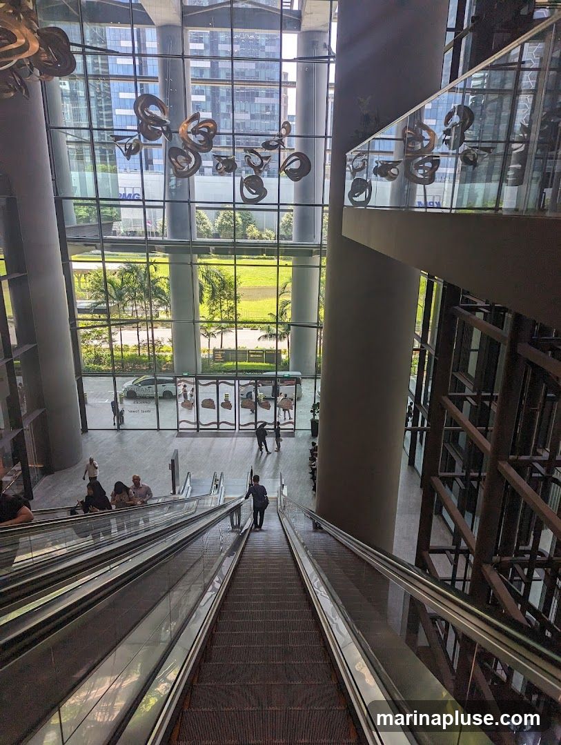 An interior view of a modern building lobby, looking down a set of escalators. Large glass windows offer a view of the exterior, including palm trees and other buildings. Decorative metal sculptures hang from the ceiling. People are seen on the escalators and in the lobby area.