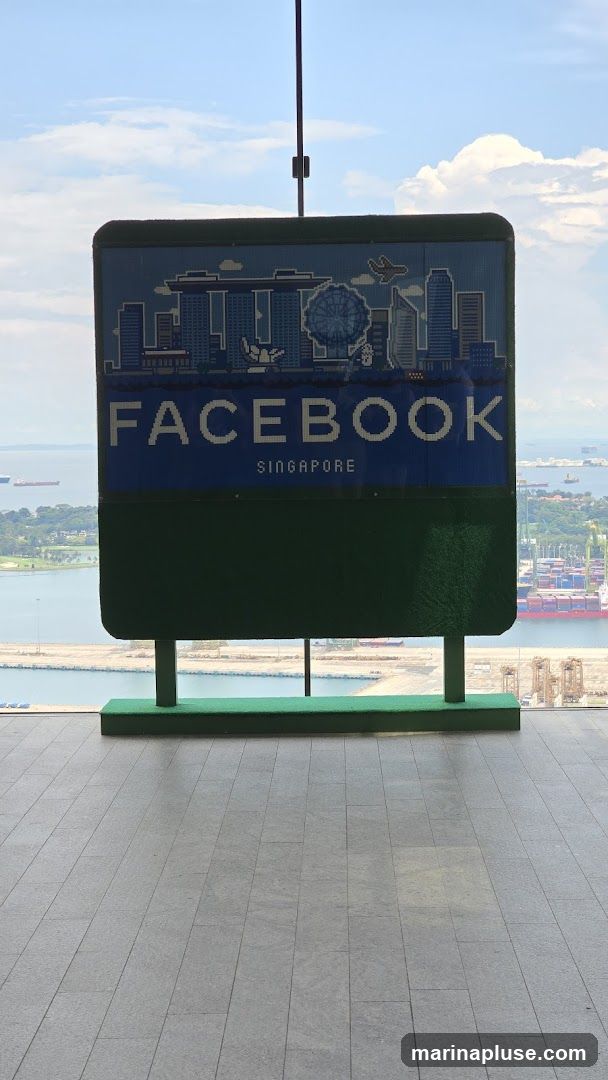 A large, pixelated sign featuring the Facebook logo and the word "SINGAPORE" below it. The sign also displays a silhouette of the Singapore skyline, including iconic buildings and the Singapore Flyer. The sign is green and blue and is mounted on a green base. The background shows a panoramic view of a harbor with ships and containers, under a bright sky with clouds.