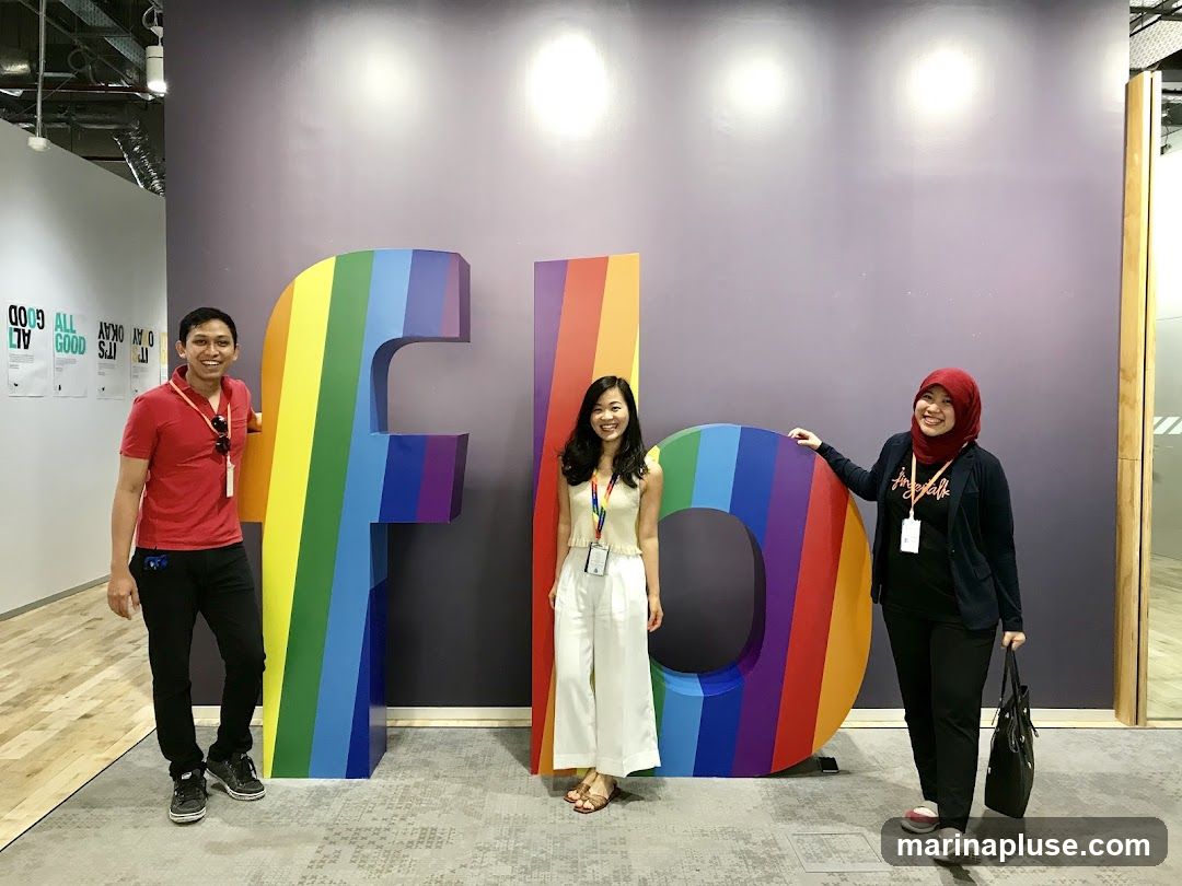 Three people are posing in front of large, colorful letters that spell out "FBO" in rainbow colors. The letters are mounted on a purple wall. The person on the left is a man in a red shirt and black pants. The person in the middle is a woman in a white top and white pants. The person on the right is a woman wearing a red hijab, a dark jacket, and black pants. There are posters on the wall to the left of the letters.