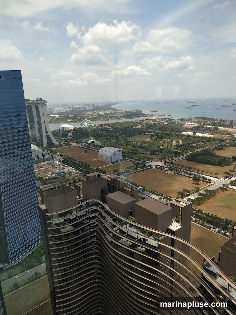An aerial view of a cityscape, likely Singapore, featuring the iconic Marina Bay Sands hotel and the Gardens by the Bay. The foreground shows the distinctive wavy architecture of a modern building. In the background, the sea is dotted with numerous cargo ships. The sky is partly cloudy.