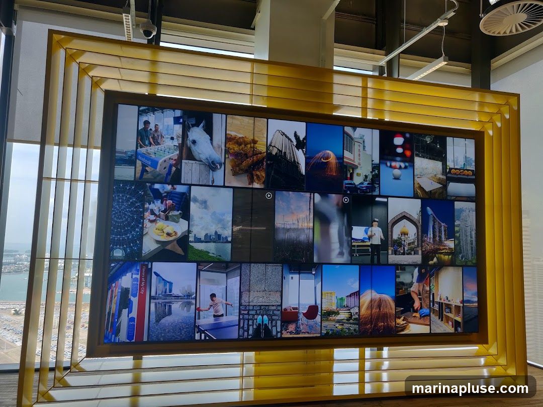 A large, multi-panel digital display mounted in a golden frame. The screen shows a collage of various images, including people playing foosball, a horse, food, cityscapes, and abstract patterns. The display is positioned indoors, with a window and blinds visible to the left, offering a view of a harbor and cityscape.