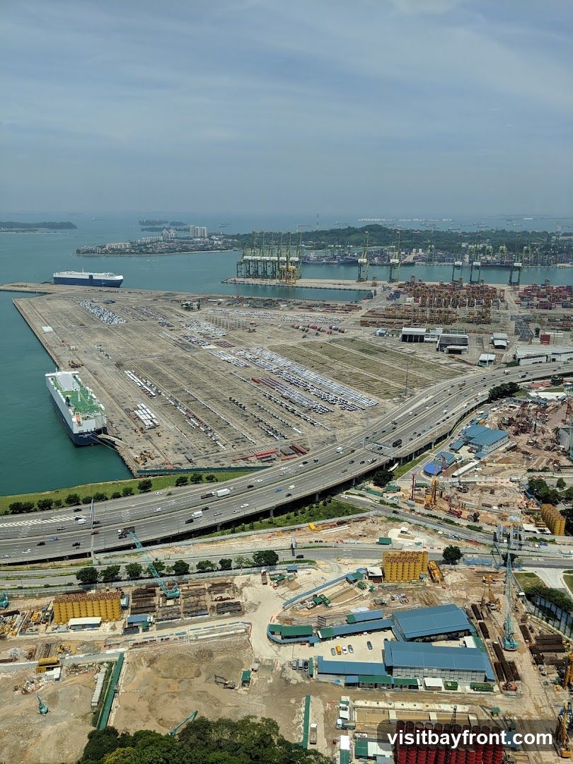 An aerial view of a busy port with numerous cargo ships, cranes, and stacked containers. A large car carrier ship is docked, and vast lots are filled with parked vehicles. A multi-lane highway runs through the scene, with traffic visible. In the foreground, a construction site is active with heavy machinery, building materials, and partially constructed structures. The sky is partly cloudy.