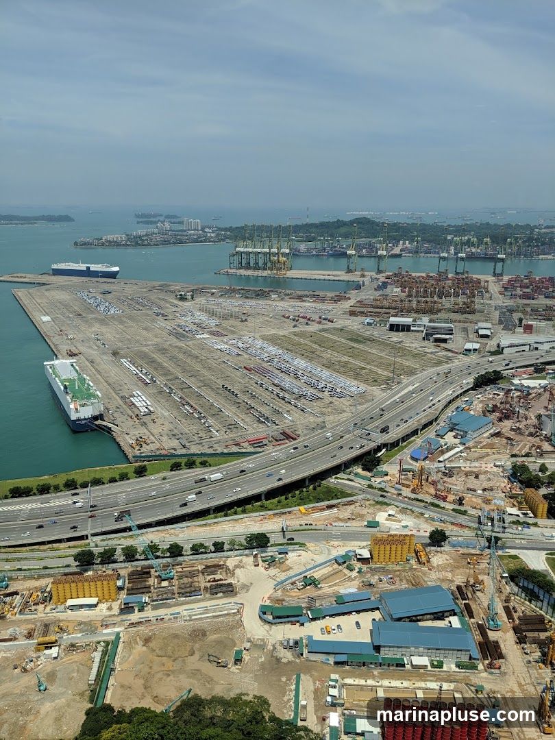 An aerial view of a busy port with numerous cargo ships, cranes, and stacked containers. A large car carrier ship is docked, and vast lots are filled with parked vehicles. A multi-lane highway runs through the scene, with traffic visible. In the foreground, a construction site is active with heavy machinery, building materials, and partially constructed structures. The sky is partly cloudy.