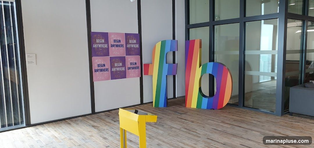Large, colorful letters spelling out "fb" in rainbow stripes stand on a wooden floor. To the left, a grid of six posters with the text "BEGIN ANYWHERE" is displayed on a white wall. A yellow stand is positioned in front of the posters. The background shows a modern interior with glass walls and doors.