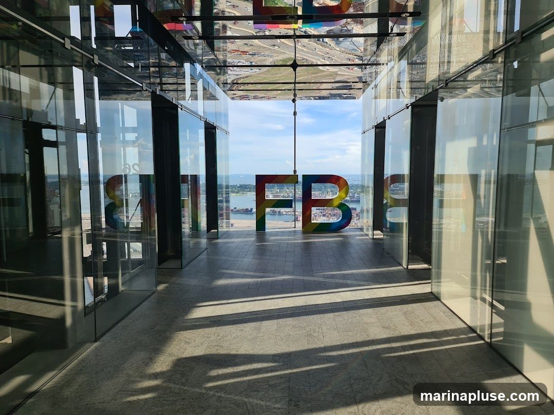 A view through a modern glass hallway towards large, colorful rainbow letters spelling out "FB". The letters are positioned in front of a panoramic city and harbor view. The hallway has a reflective ceiling and glass walls, creating reflections of the interior and the exterior scene. Sunlight casts long shadows across the tiled floor.