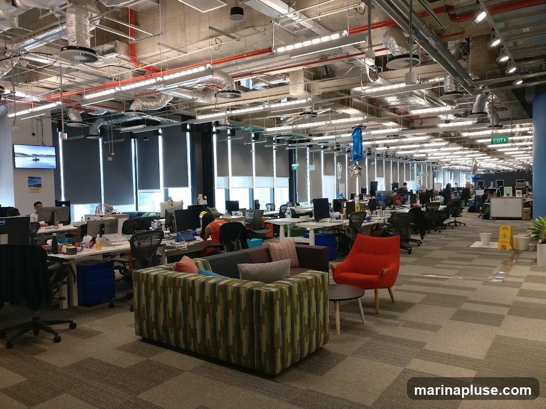 An expansive, modern office space with rows of desks, computers, and office chairs. The ceiling features exposed ductwork and lighting. In the foreground, a colorful sofa and an orange armchair create a lounge area. The background shows more workstations and an exit sign.