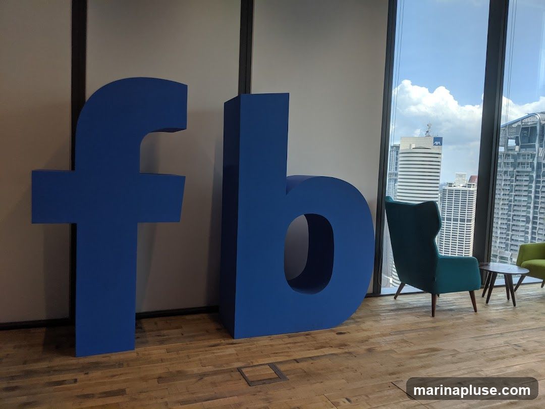 Large blue letters spelling "fb" are displayed in an office setting. The letters are three-dimensional and stand on a wooden floor. Behind the letters, a large window offers a view of a city skyline with several tall buildings. Two armchairs, one teal and one green, are placed near the window, along with a small round coffee table. The overall impression is of a modern and spacious office environment, likely a reception or common area.