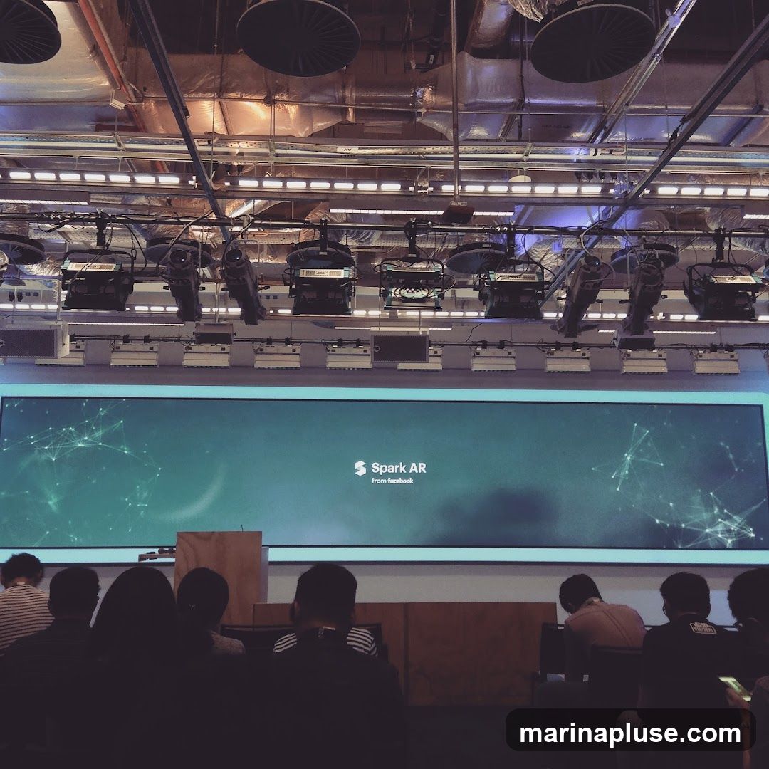 An audience watches a presentation on a large screen displaying "Spark AR from Facebook." The screen is set against a backdrop of stage lighting and equipment.