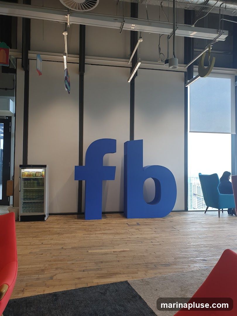 A large, blue, three-dimensional "fb" logo is displayed prominently in a modern office space. To the left of the logo is a glass-fronted refrigerator stocked with beverages. In the background, there are large windows offering a view of a city skyline, and several colorful chairs are visible. The floor is made of light-colored wood planks.