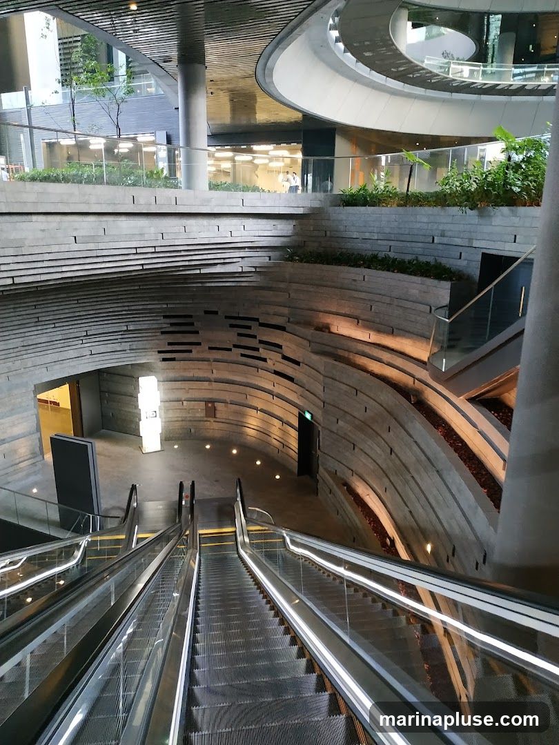 An interior view of a modern building featuring escalators descending into a multi-level space. The architecture includes curved concrete walls with layered stone accents, glass railings, and integrated lighting. Greenery is visible on upper levels, adding a natural element to the contemporary design.