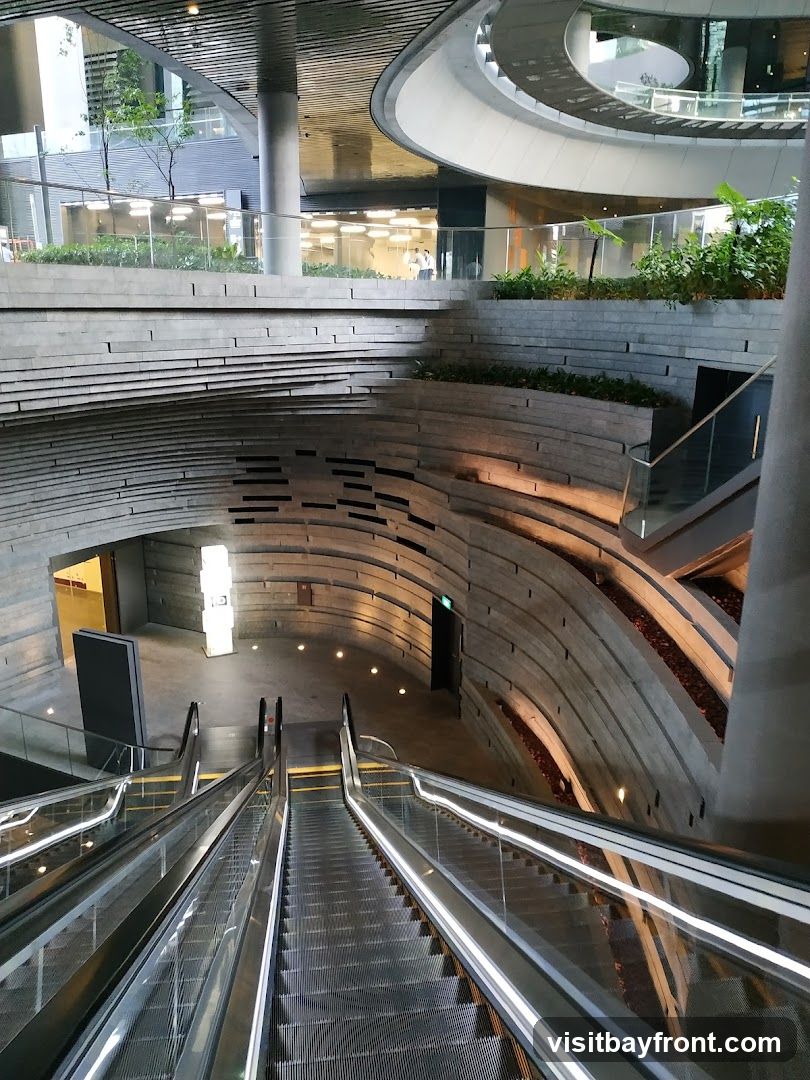 An interior view of a modern building featuring escalators descending into a multi-level space. The architecture includes curved concrete walls with layered stone accents, glass railings, and integrated lighting. Greenery is visible on upper levels, adding a natural element to the contemporary design.