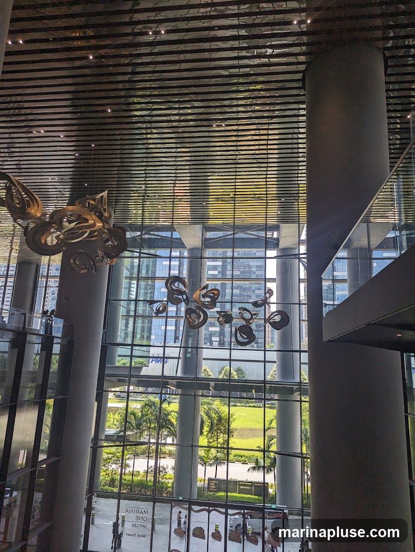 An interior view of a modern atrium with a high, reflective ceiling and large glass windows overlooking an outdoor green space and city buildings. Two large, abstract metal sculptures hang from the ceiling. Large cylindrical columns support the structure.