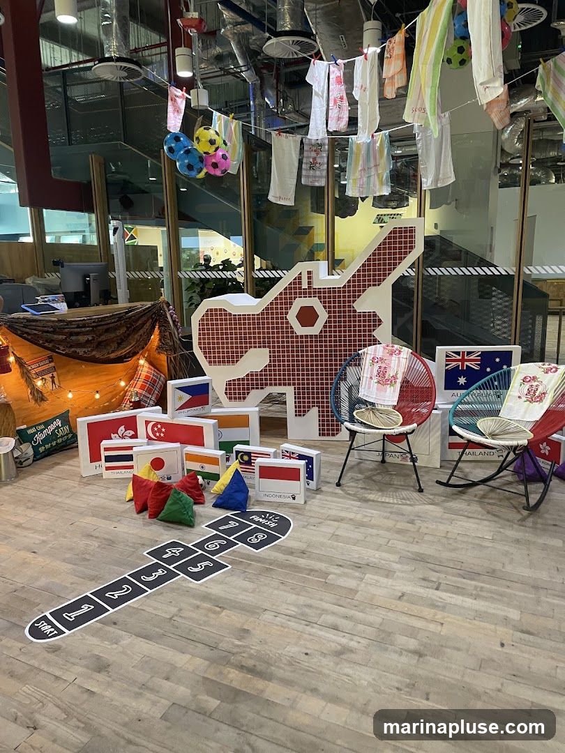 A festive indoor display features flags of various countries, including Hong Kong, Singapore, Thailand, India, Malaysia, and Indonesia. A large, stylized red and white animal cutout stands prominently. Clothes are hung on a line above, with colorful soccer balls. Two chairs, one rocking, are placed near the cutout, adorned with decorative cloths. A hopscotch grid is marked on the wooden floor.