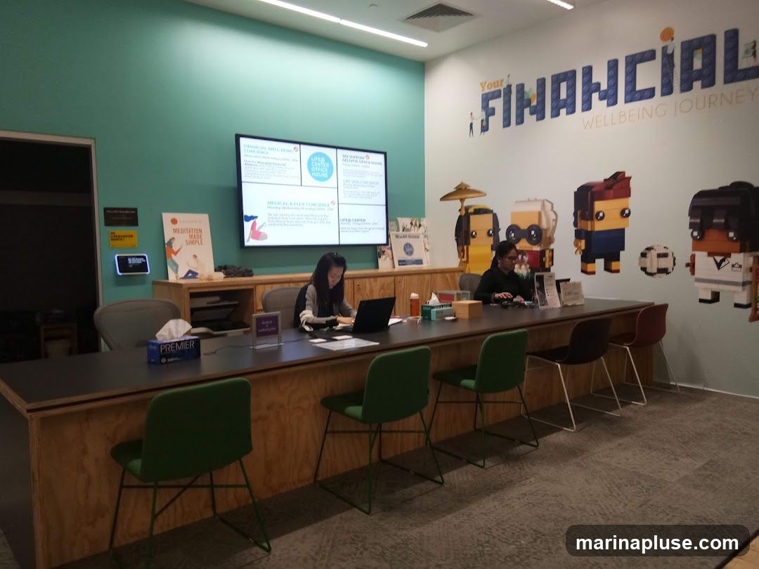 An interior view of a modern office space with a long counter where two employees are working. The wall behind them is decorated with colorful Lego-like figures and the text "Your FINANCIAL WELLBEING JOURNEY". A large screen displays information, and various posters and brochures are visible.