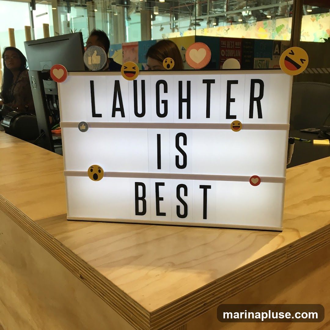 A white lightbox with black lettering displays the quote "LAUGHTER IS BEST". The lightbox is decorated with various emoji stickers, including laughing faces, a heart, and a thumbs-up. The lightbox sits on a wooden counter in what appears to be an office or communal space. In the background, people are visible, along with office equipment and colorful wall decorations.