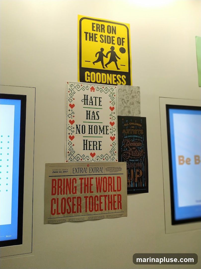 A wall with several posters and two digital screens. One poster is yellow with black text and silhouettes of two children. Another poster has a cross-stitch design with the text "HATE HAS NO HOME HERE". A newspaper-style poster reads "BRING THE WORLD CLOSER TOGETHER". A dark poster has text about children and artists. One digital screen displays blue dots, and the other has orange text.