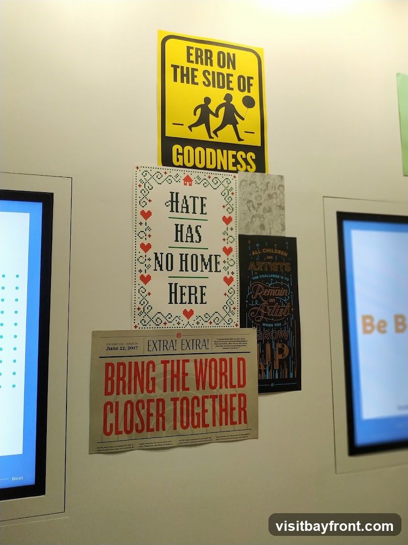 A wall with several posters and two digital screens. One poster is yellow with black text and silhouettes of two children. Another poster has a cross-stitch design with the text "HATE HAS NO HOME HERE". A newspaper-style poster reads "BRING THE WORLD CLOSER TOGETHER". A dark poster has text about children and artists. One digital screen displays blue dots, and the other has orange text.
