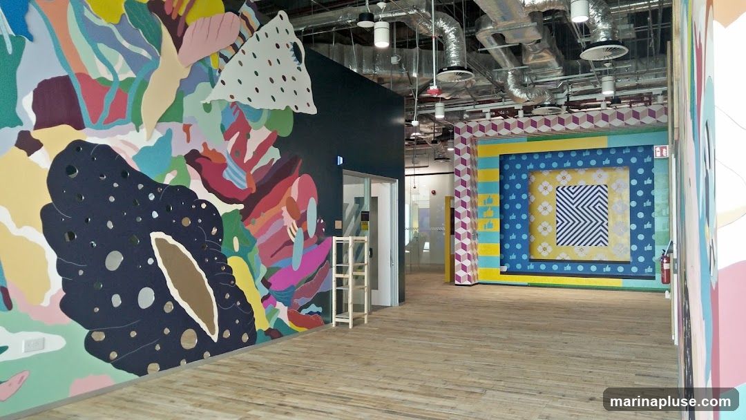 The image shows a brightly decorated interior space with large, abstract, colorful murals on the walls. One wall features a dark blue background with a large, organic shape in shades of dark blue and brown, with circular cutouts. Another wall has a vibrant, abstract design with organic shapes in various colors like pink, purple, yellow, and green. In the background, there is a wall with a large, framed geometric pattern in blue, yellow, and white, with repeating square motifs and a central zigzag design. The floor is made of light-colored wooden planks. The ceiling has exposed industrial elements like ventilation ducts and lighting fixtures. There is a doorway with a glass door and a small wooden shelf unit next to it. A fire extinguisher is visible on the right side of the image.