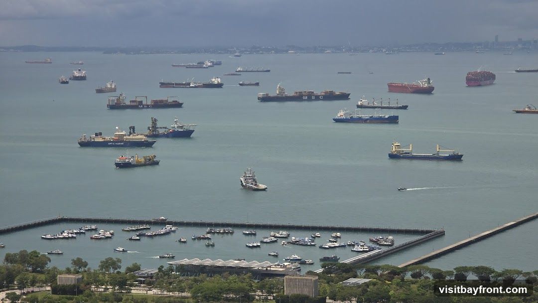 An aerial view of a busy harbor filled with numerous cargo ships and smaller vessels. The ships are of various sizes and types, including container ships, tankers, and tugboats, anchored or moving in the water. In the foreground, there is a pier with many smaller boats docked, and beyond that, a lush green coastline with some buildings and trees. The sky is overcast, casting a muted light over the scene.