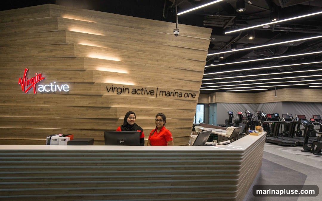 Two women stand behind a reception desk at a Virgin Active gym. The Virgin Active logo is visible on the wall behind them, along with the text "virgin active | marina one". In the background, rows of treadmills and other exercise equipment are visible.