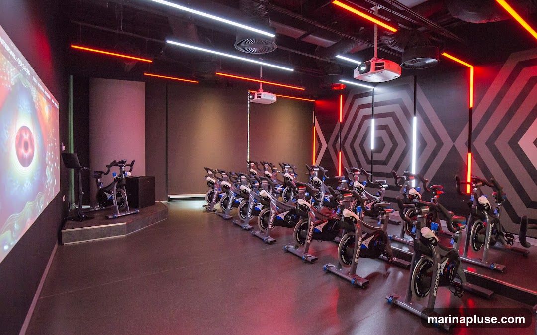 An indoor cycling studio with rows of stationary bikes. A large screen is projected on the left wall, displaying abstract visuals. The room is lit with red and white LED strip lights, and a projector is mounted on the ceiling. The walls have a geometric hexagonal pattern.