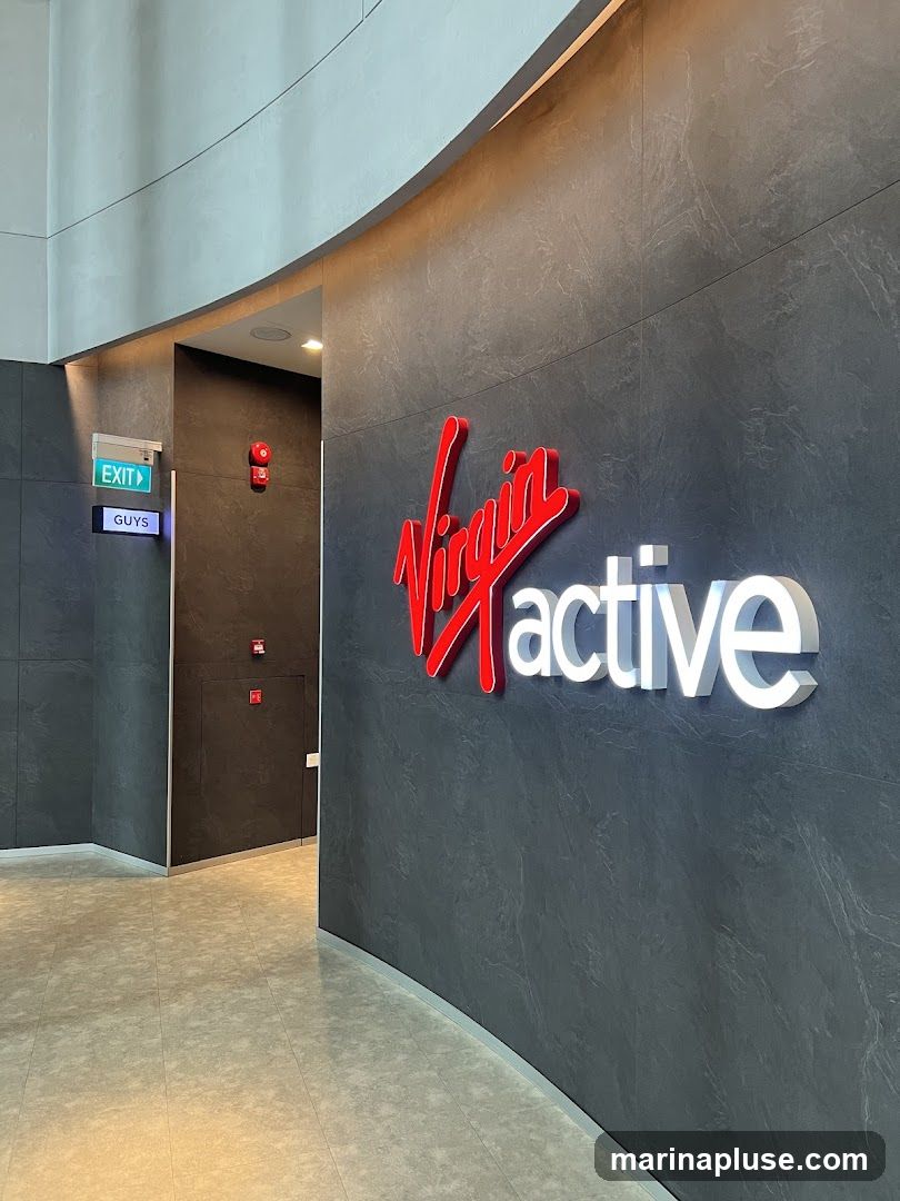 A large red and white Virgin Active logo is displayed on a dark gray textured wall. To the left, an illuminated EXIT sign is visible above a sign that reads GUYS. The floor is tiled with a light beige pattern.