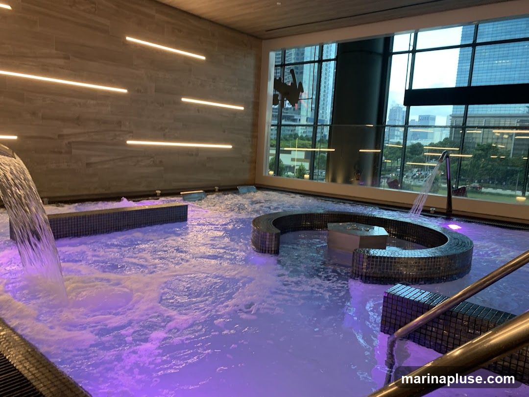 A luxurious indoor spa features a large pool with bubbling water illuminated by purple lights. Waterfalls cascade into the pool from the left side, and jets create a swirling effect. A curved, tiled seating area with a central platform is submerged in the water. The pool area has modern, tiled walls with horizontal strip lighting. A large window offers a panoramic view of a city skyline with tall buildings and greenery. Metal railings and steps provide access to the pool.