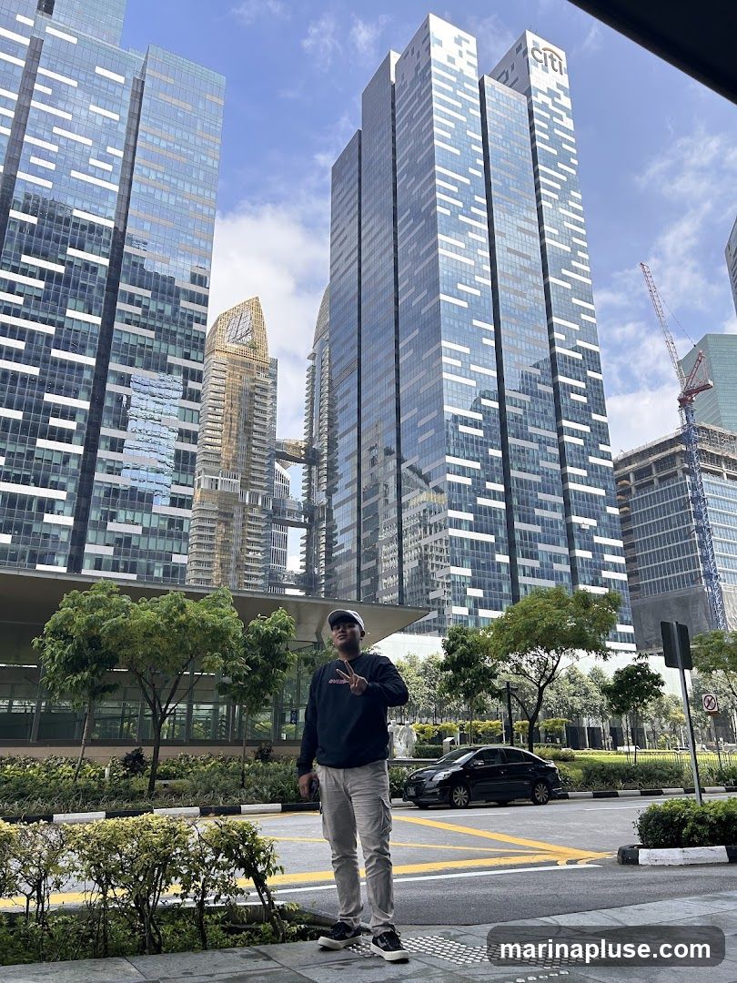 A man stands on a sidewalk in front of several modern skyscrapers. The buildings are tall and made of glass and steel, with a distinctive pattern of white lines on their facades. One of the buildings has a prominent "citi" logo at the top. The sky is blue with some clouds. There are trees and landscaping along the sidewalk, and a black car is parked on the street. The man is wearing a dark sweatshirt, light-colored cargo pants, a baseball cap, and sneakers. He is holding a phone in his left hand and making a peace sign with his right hand.