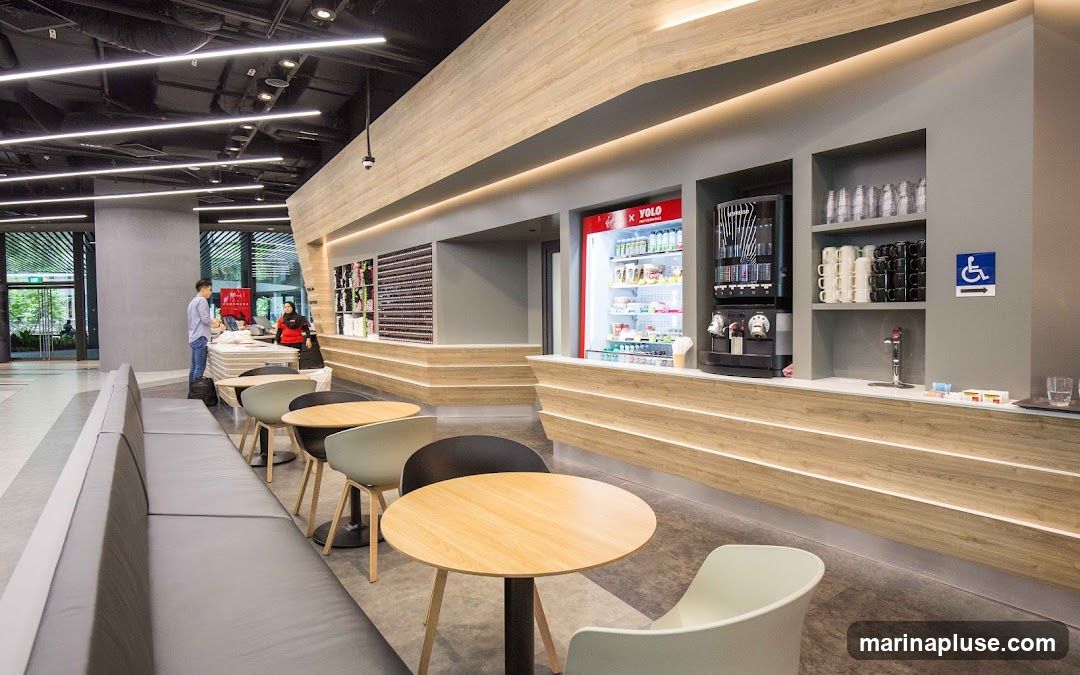 A contemporary cafe with a long grey sofa seating area, round wooden tables, and chairs. The counter features a refrigerated display case with drinks, a coffee machine, and shelves stocked with cups and mugs. A sign indicating accessibility is visible on the wall.