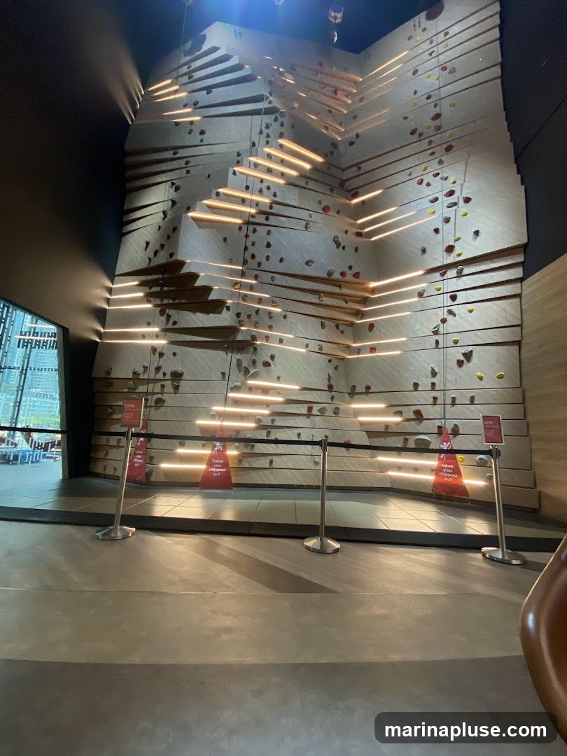 An indoor climbing wall with numerous holds and illuminated horizontal ledges. The wall has a modern, angular design. Stanchions with red triangular signs are placed in front of the wall.