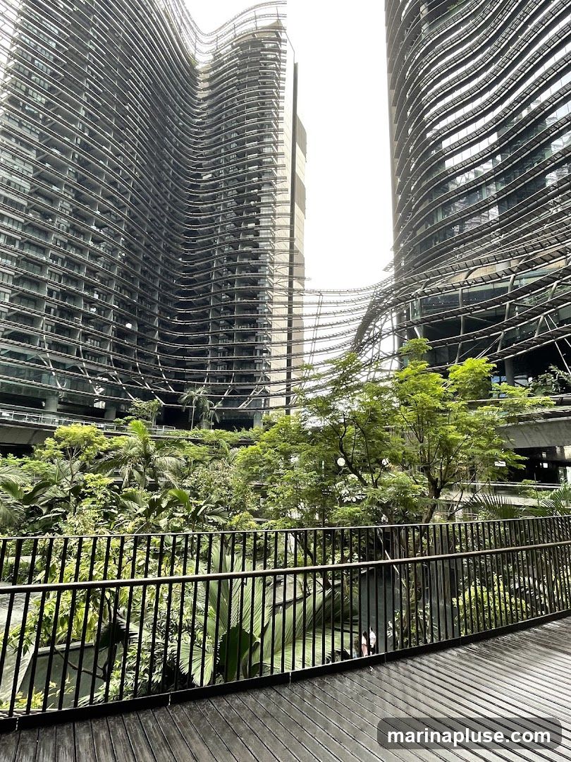 A view of two futuristic, wave-like skyscrapers with a central atrium filled with lush tropical plants. A wooden walkway with a black metal railing is in the foreground, overlooking the greenery and a body of water.