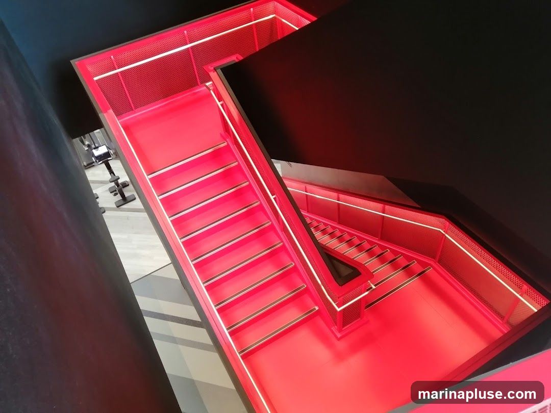 A high-angle view of a modern staircase with bright red steps and railings, illuminated by white LED lights along the edges. The surrounding walls are dark, creating a stark contrast with the vibrant staircase. In the background, a gym area with exercise equipment is partially visible.