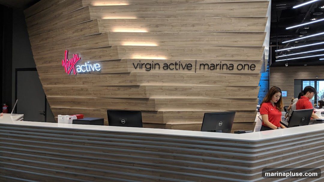 A reception desk area with staff members wearing red shirts. The wall behind the desk features a prominent "Virgin Active" logo and the text "virgin active | marina one". The reception desk is modern with a white countertop and a wood-paneled base. There are computer monitors on the desk.